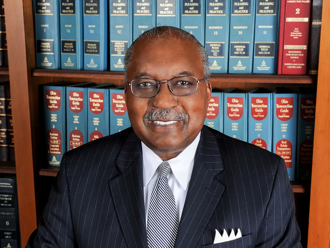 This portrait of Ben Espy shows a sophisticated Black man with a friendly smile. He wears glasses and a pin-striped suit and stands in front of shelves lined with law books.