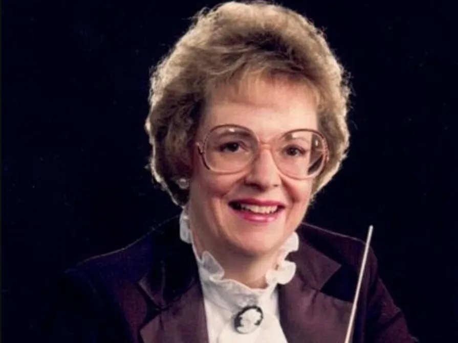 Carol Clark poses for a portrait holding a conductor's baton and dressed for an orchestra. She's smiling, has 1980s-style rounded frame glasses, pearl earrings and short blonde hair attractively poofed.