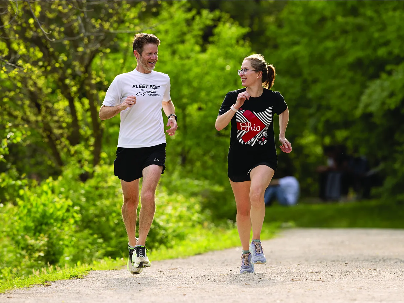A woman and man talk as they run down a bicycle trail surrounded by trees and lush vegetation.