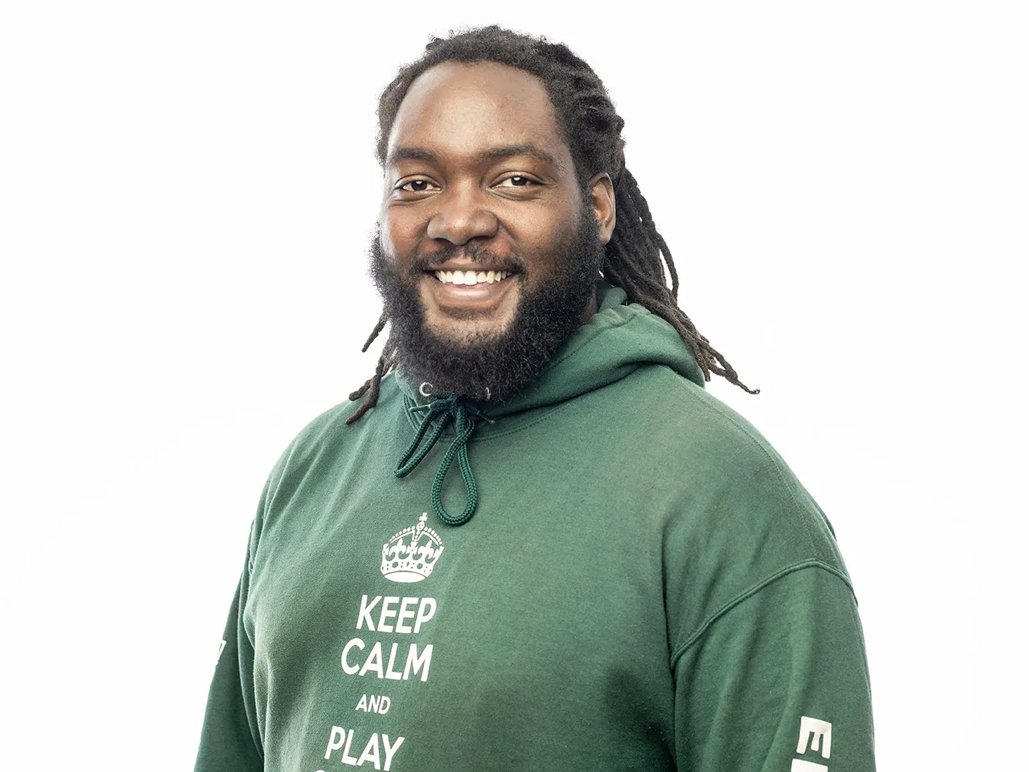 In this portrait of Ernest Levert Jr., he poses with a friendly smile. His sweatshort says "Keep calm and play chess" and has a beard and thin dreads pulled back