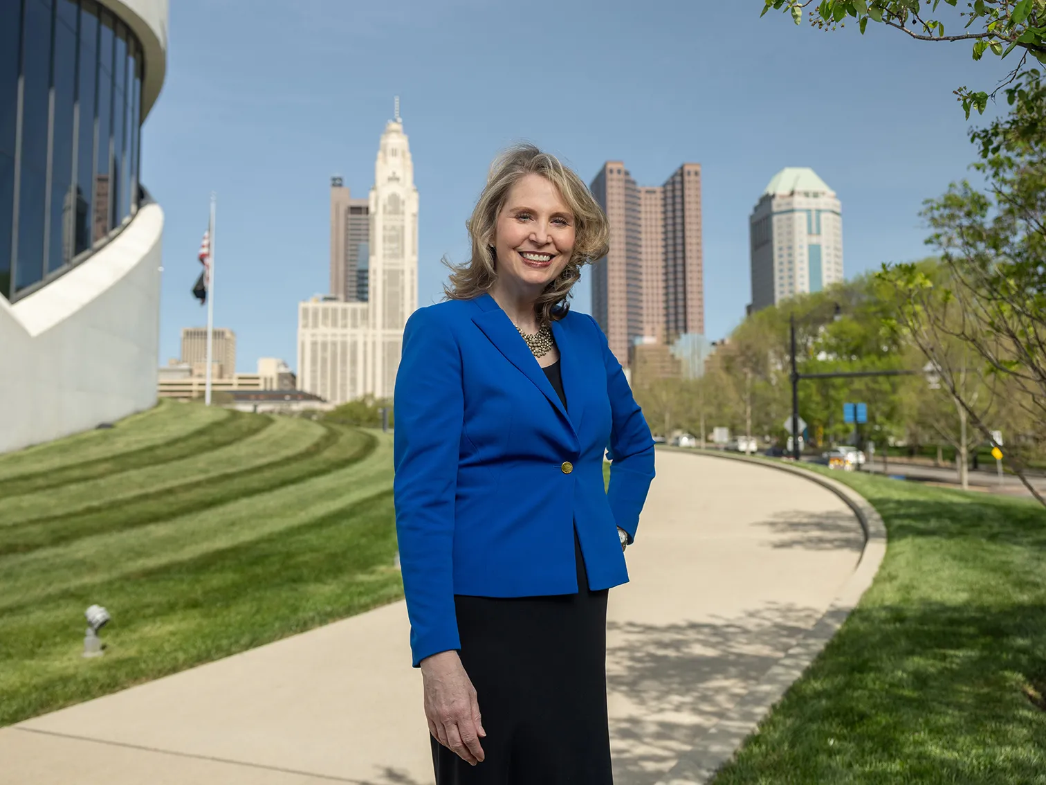 Amy Edwards Taylor smiles and stands with one hand on her hip as she poses for this photo. Behind her is the edge of the Veterans Memorial Museum and farther away, downtown Columbus. She has shoulder length blond hair, wears a business suit and smiles like she's genuinely happy to see you.
