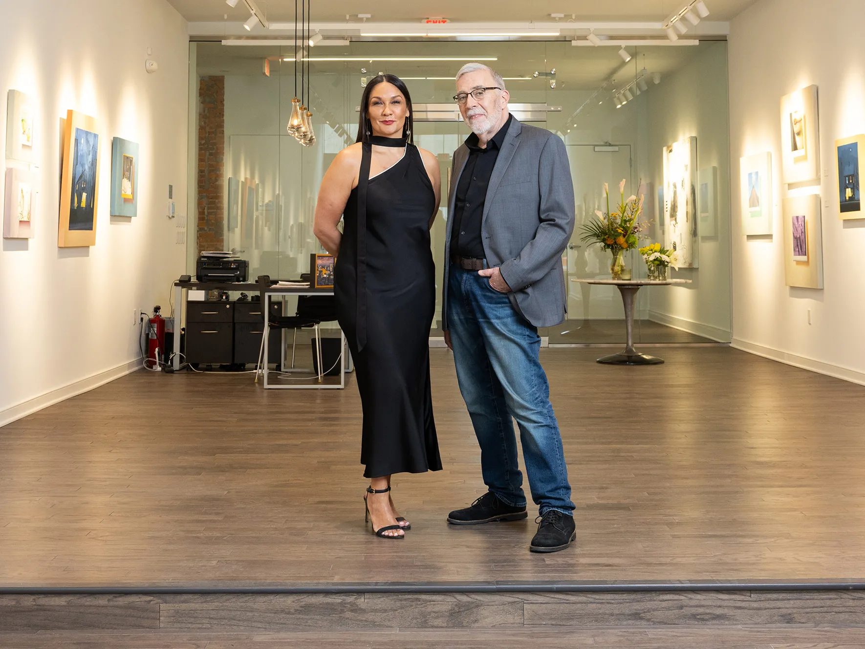 Michelle Brandt wears a black dress and Duff Lindsay wears jeans and a sports coat in a new art gallery. They seem elegant and thoughtful as they pose standing next to each other and slightly smiling.
