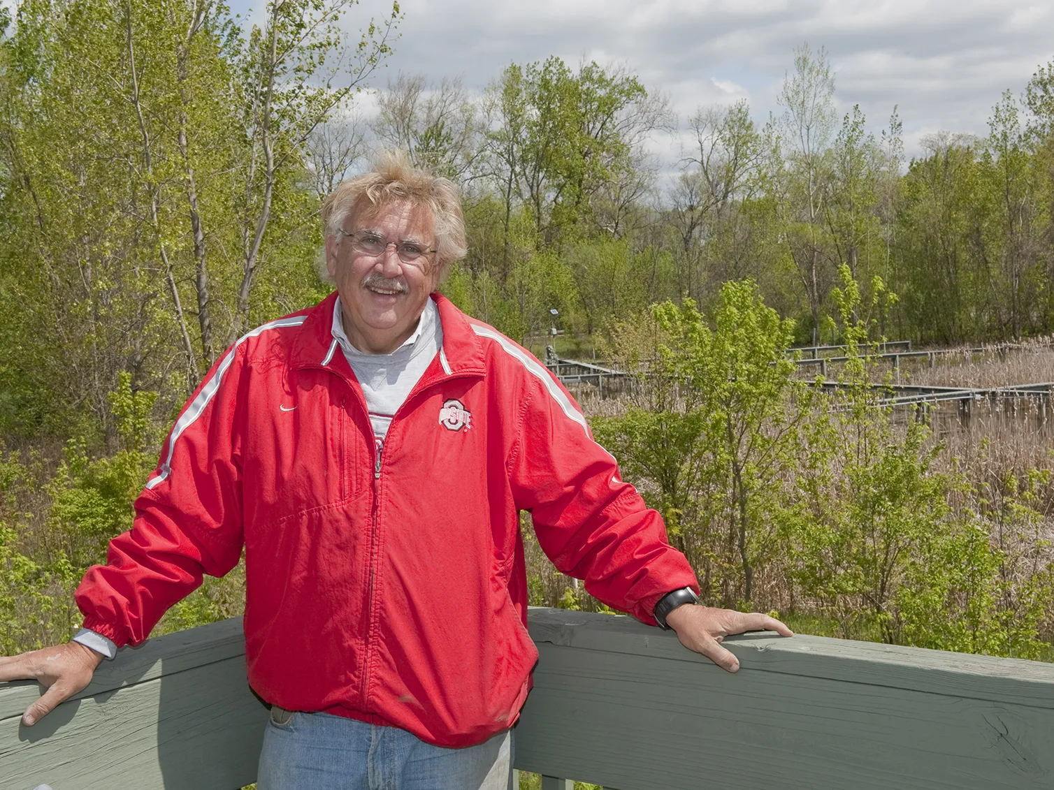 This portrait shows Bill Mitsch, a middle-age white man with a friendly smile. He's posing in front of the Olentangy River Wetland Research Park, which has raised wooden walkways over wetlands grasses inside a ring of trees.