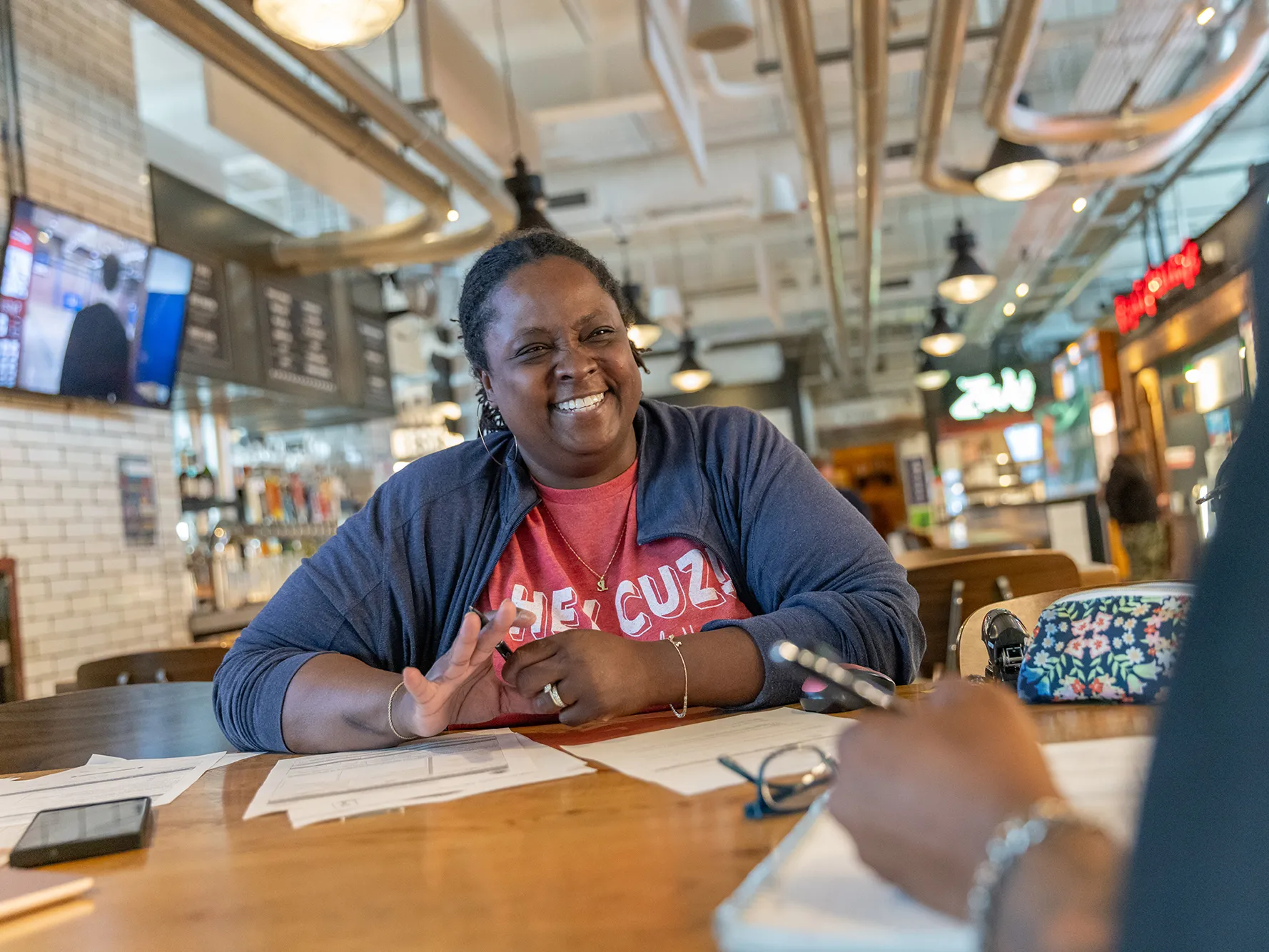 Sadaya "Daisy" Lewis sits at a hightop table to onboard an employee. This food hall is where her popular Modern Southern Table restaurant is located. There is exposed, shiny ductwork, white brick walls and a clean, modern feel.