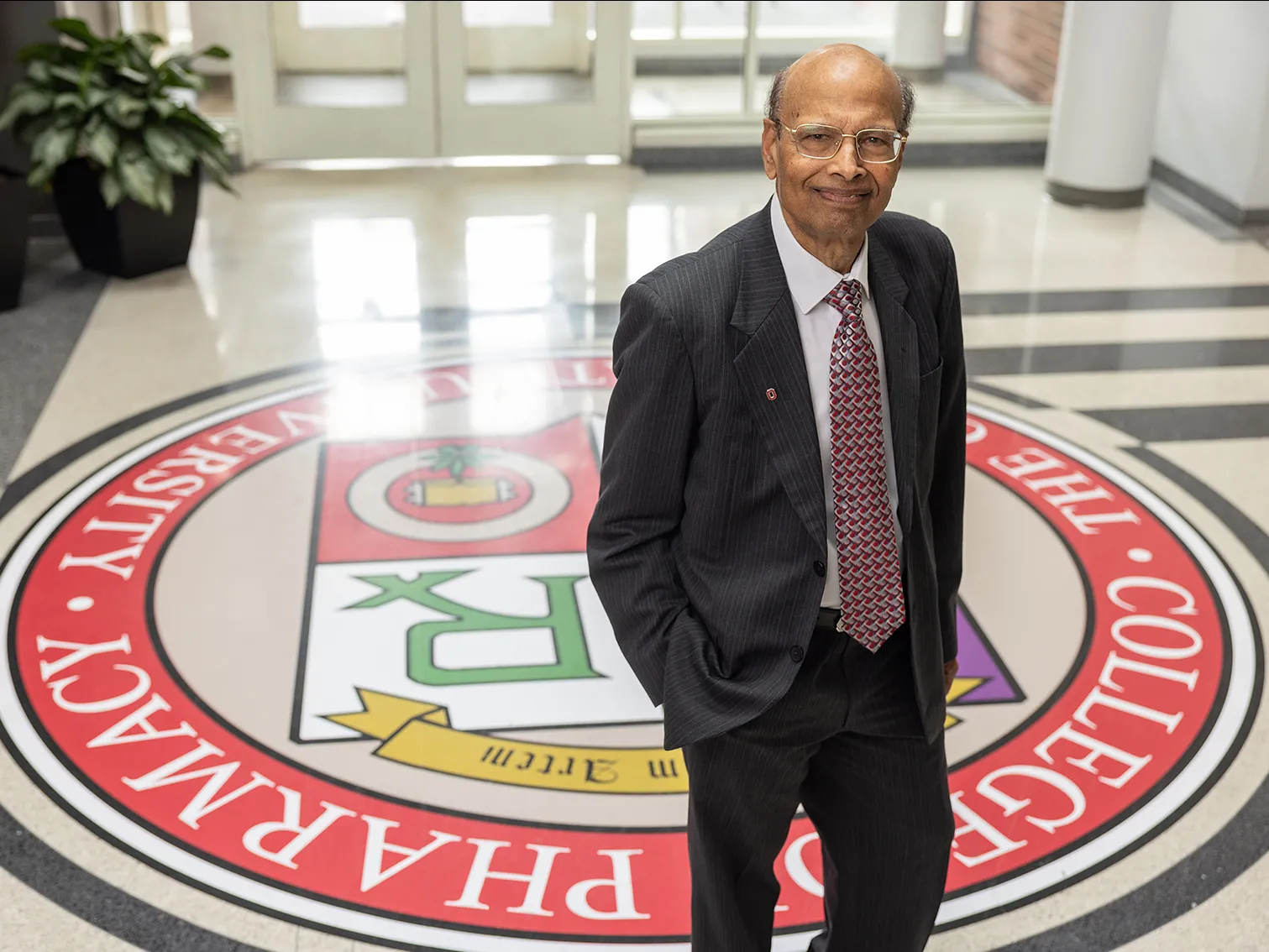 Milap Nahata, an Indian man, poses for a portrait in the lobby of the pharmacy college at Ohio State. Its colorful insignia is painted on the floor, adding an interesting background. Nahata looks calm, confident and friendly.