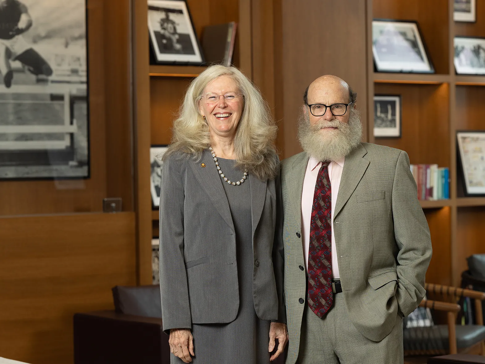 Gay Miller and Joseph Rosenblatt are a white couple who are both dressed in suits. She has long, pretty hair and is laughing. He has a lush beard and glasses. They are in the lobby of a university building. Around them are Ohio State memorabilia on wooden shelves and chairs.
