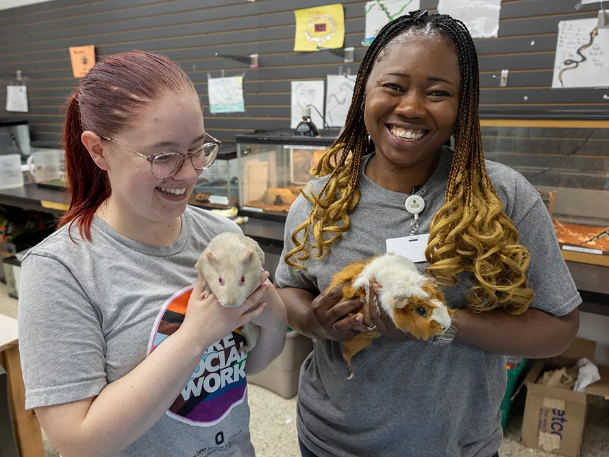 A young social worker and a graduate student from Ohio State smile as they hold guinea pigs in a room at Buckeye Ranch where therapy pets live