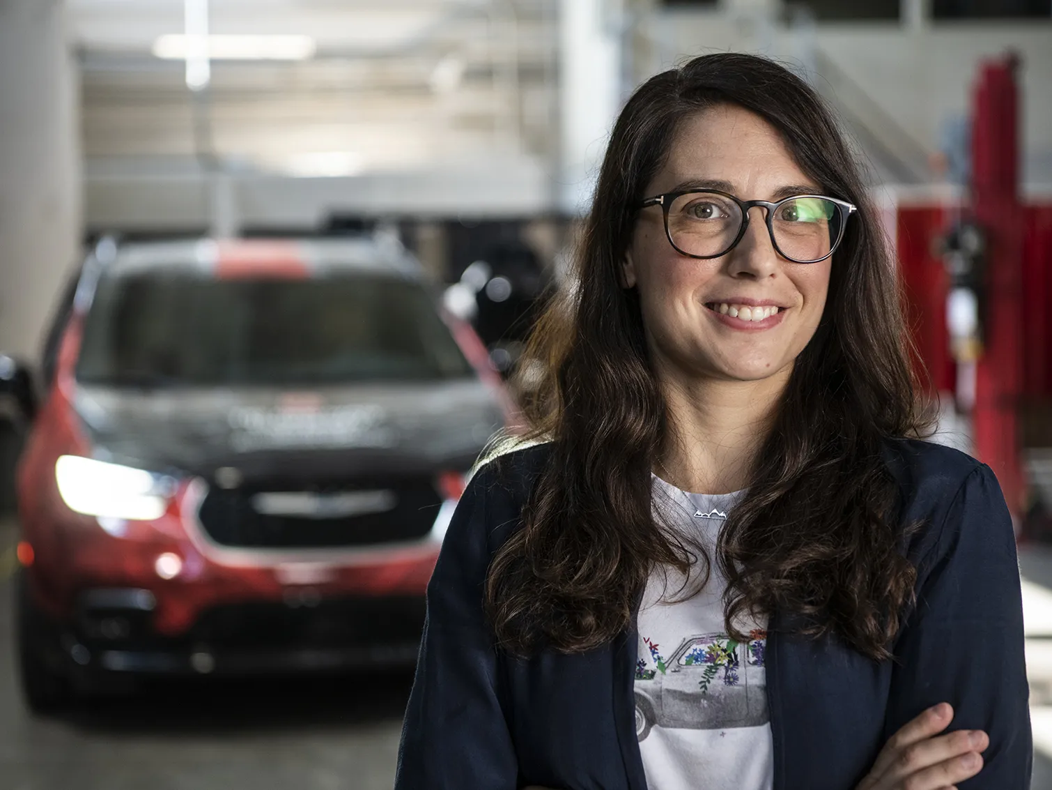 Stephanie Stockar, a white woman with glasses and long brown hair, smiles for a portrait in the automobile laboratory where she works, she wears T-shirt and sweater. Behind her is an electric car.