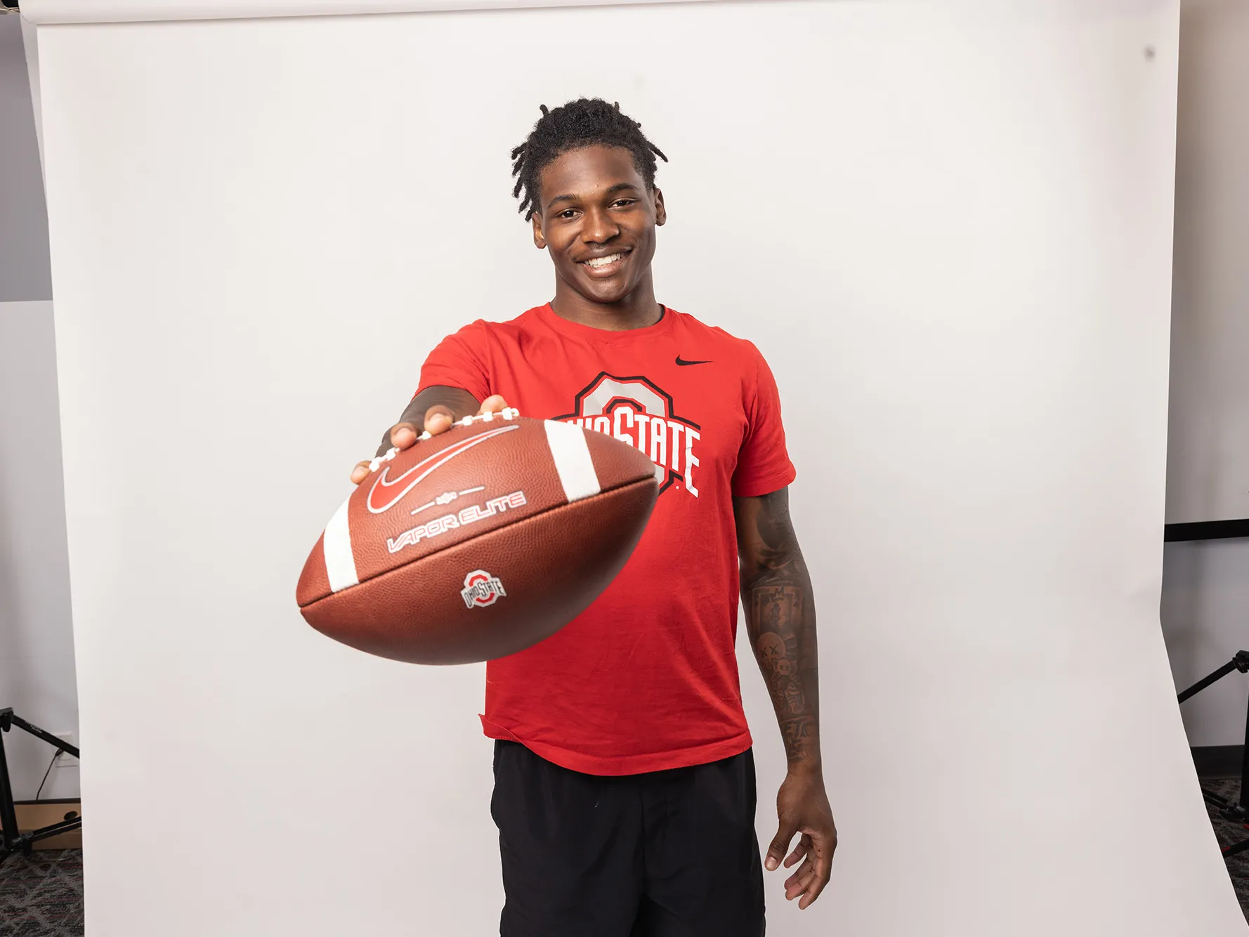 Ohio State football player Jeremiah Smith poses for a photo holding out a football. He&#039;s an attractive young black man, smiling with his hair pulled up in a ponytail and tattoos on his arms.