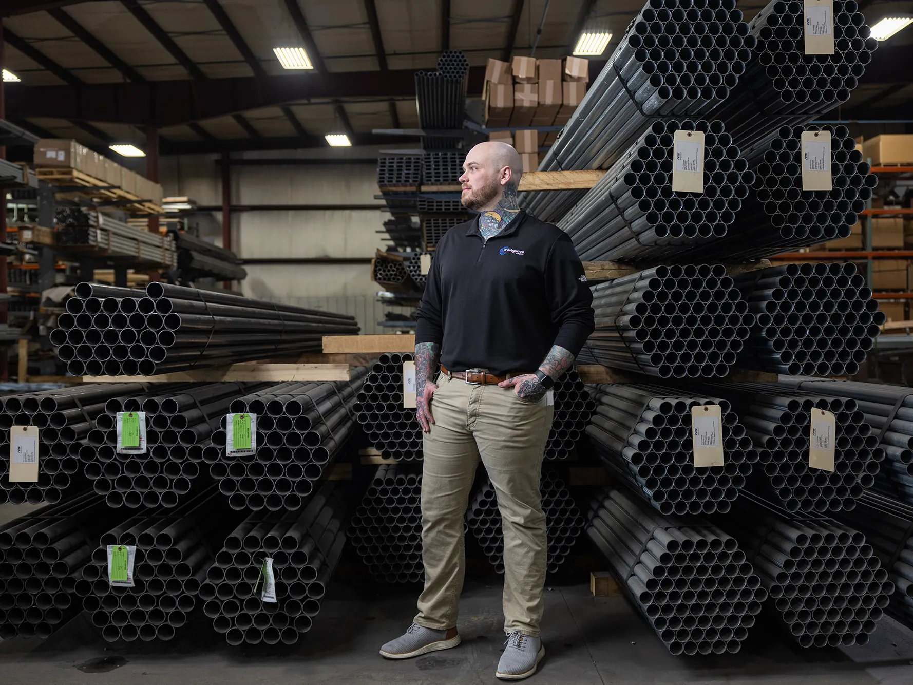 Zachary Ernest, a tall white man with shaved head, neat beard and colorful tattoos on his neck and forearms, is pleased by what he sees on the factory floor of his employer, Ashland Conveyor Products. He stands in front of bundles of about 50 metal pipes each, stacked on shelving. 