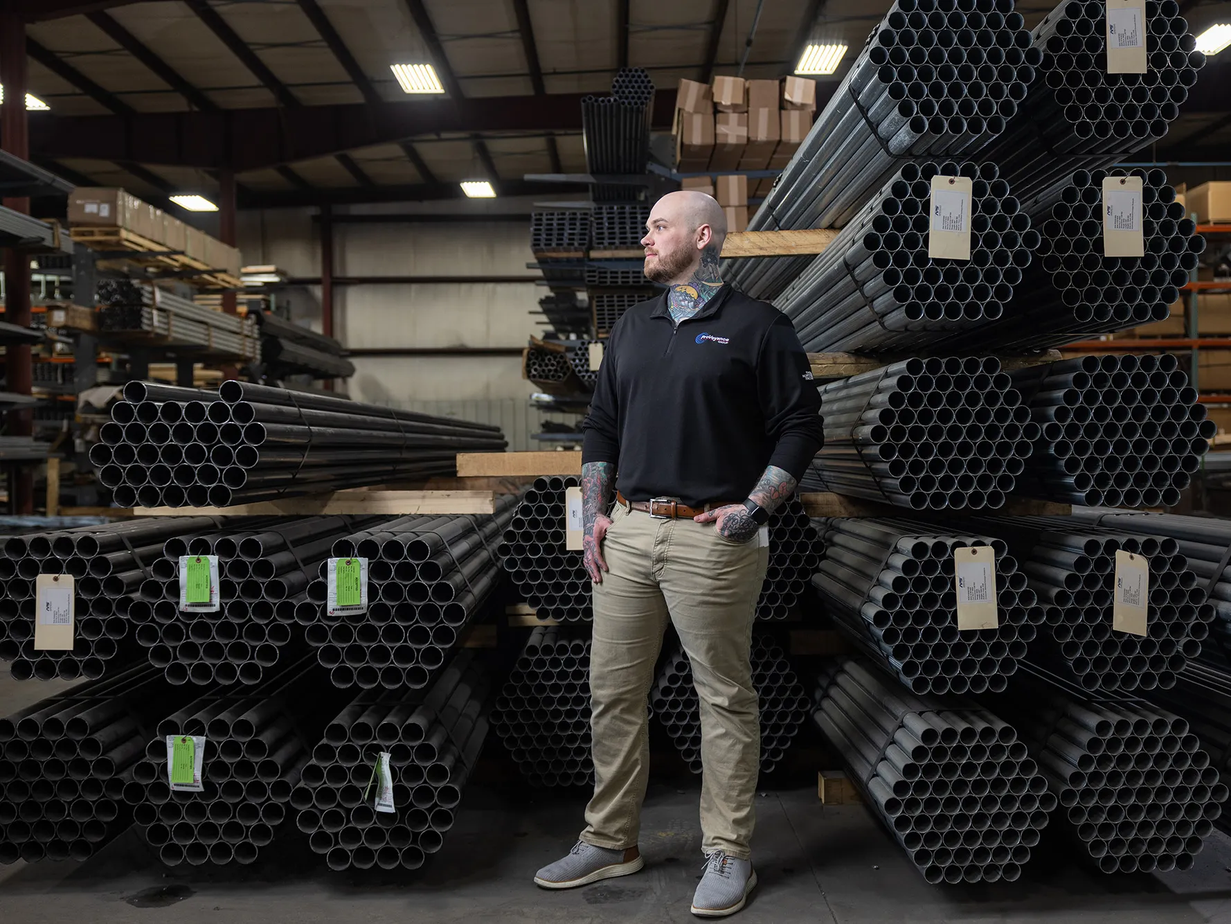 Zachary Ernest, a tall white man with shaved head, neat beard and colorful tattoos on his neck and forearms, is pleased by what he sees on the factory  floor of his employer, Ashland Conveyor Products. He stands in front of bundles of about 50 metal pipes each, stacked on shelving. 