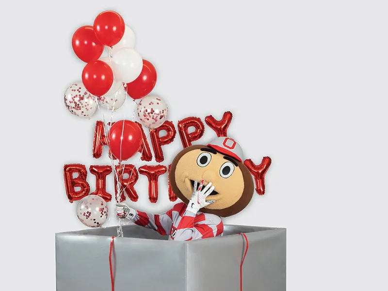 Brutus Buckeye puts a hand to his mouth as he giggles while rising from inside a giant gift-wrapped box.