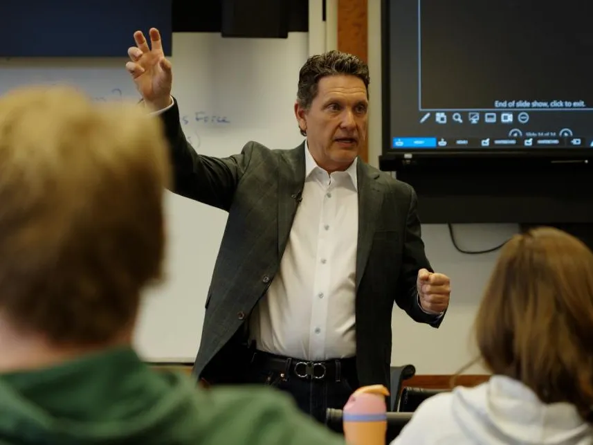 Ohio State alumnus Jeff Prestel talks to students during a class at Fisher College of Business. He stands in front of them, arm raised as he gestures to make a point. He seems professional and focused.