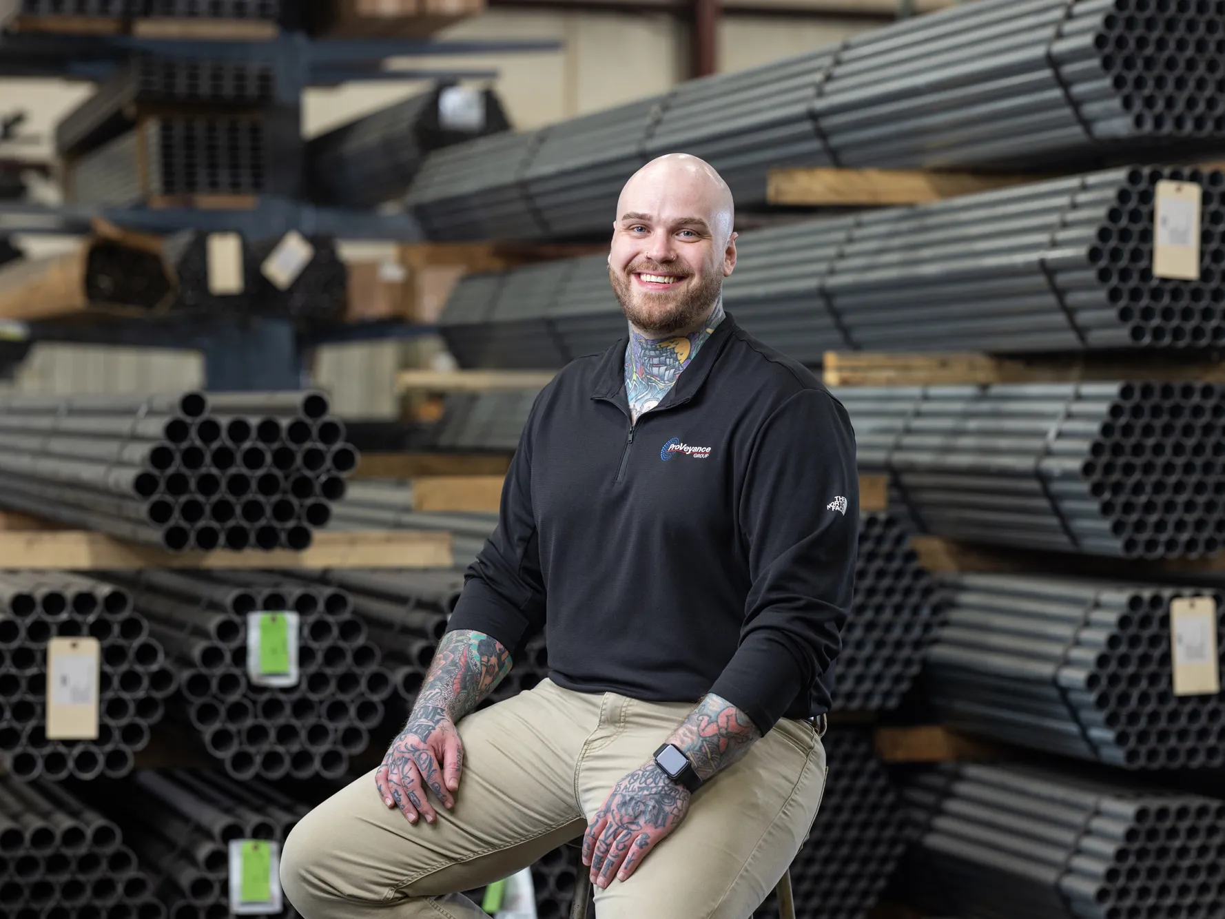 Zachary Ernest, a tall white man with shaved head, neat beard and colorful tattoos on his neck and forearms, smiles as he sits on a stool on the factory floor of Ashland Conveyor Products. He is in front of bundles of about 50 metal pipes each, stacked on shelving. 