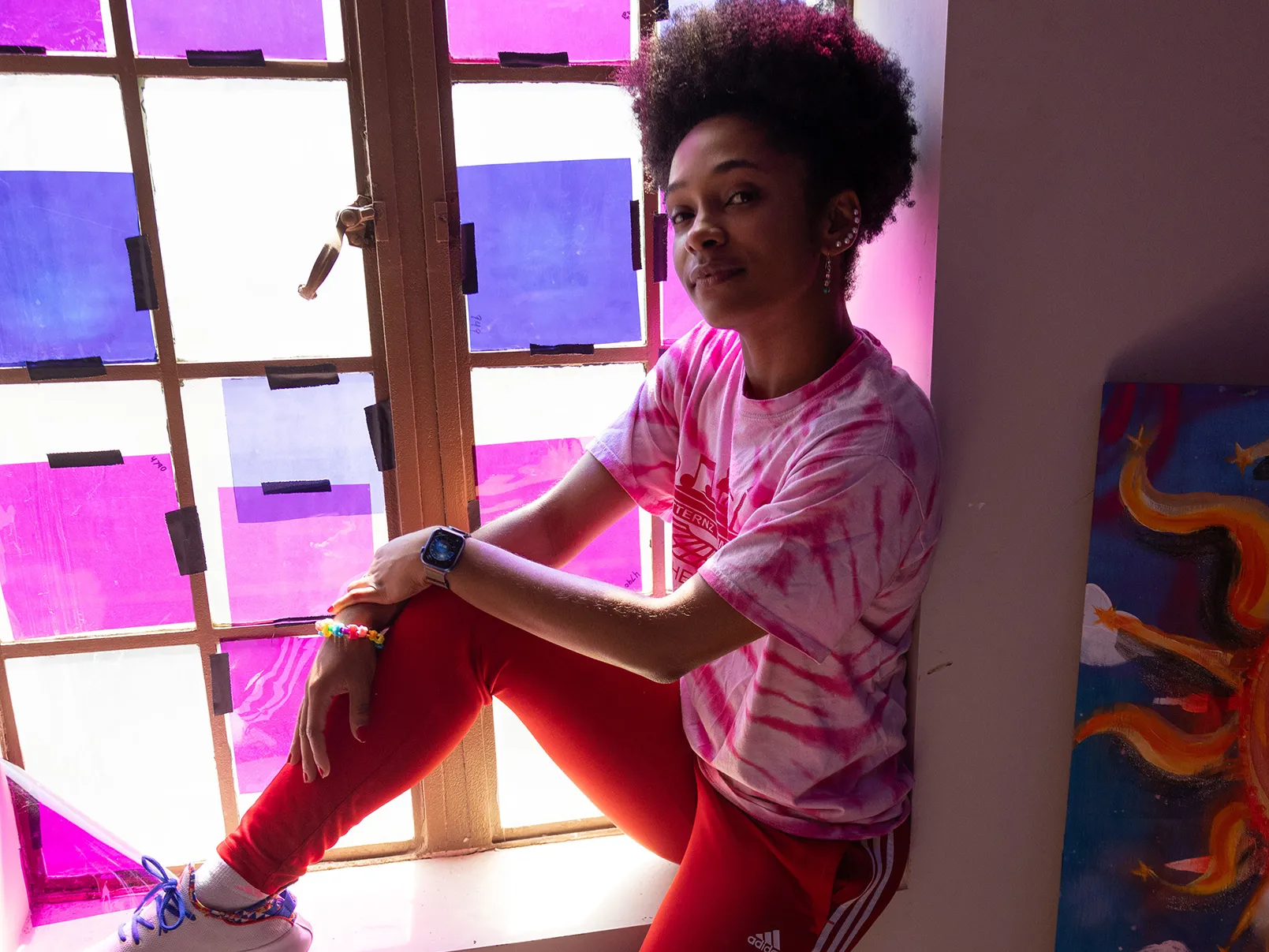 Brianna Rhodes, a pretty, young black woman, sits on a window ledge inside a dim room. Colored paper taped over the window shines colored light onto her. She&#039;s smiling with her eyes and wearing casual clothes that she can dance in. Her hair is tight curls on top of her head and she has a line of earrings up the ear the photo shows. 