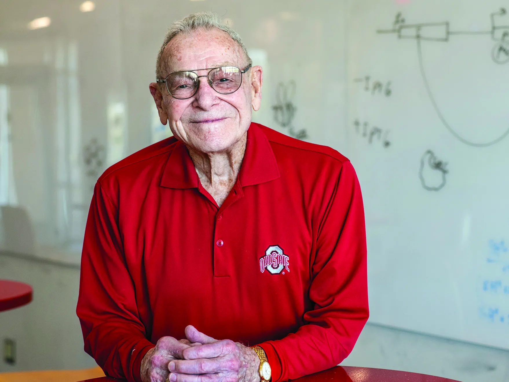 Merle Lashey smiles with closed lips, but not less enthusiasm than a big grin, in a classroom on the Marion campus. He is an older white man wearing a scarlet Ohio State golf shirt, and behind him is a white board with hand-drawn diagrams.