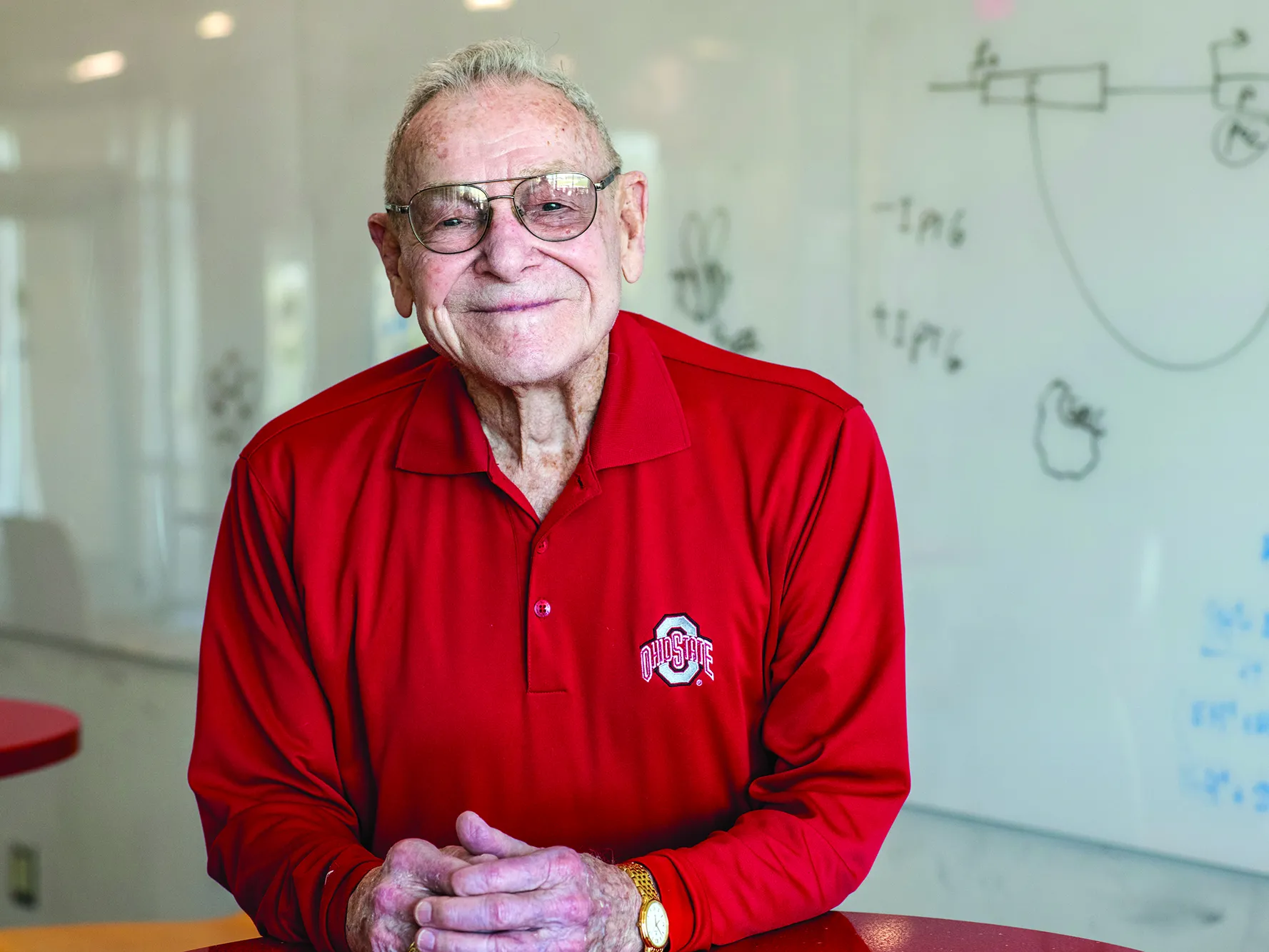 Merle Lashey smiles with closed lips, but not less enthusiasm than a big grin, in a classroom on the Marion campus. He is an older white man wearing a scarlet Ohio State golf shirt, and behind him is a white board with hand-drawn diagrams.