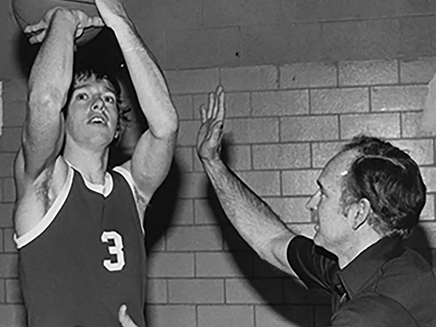 In an older black and white photo, Gene Millard instructs a young man who's holding a basketball as if he's ready to shoot a basket. The coach is either playing defense or correcting the young man's form. He is slightly crouched and has one hand higher in the air. Neither is grinning, but they both look as if they're enjoying themselves.