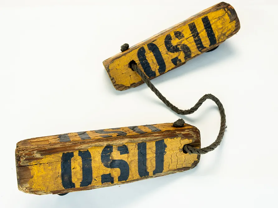 Two weathered, long triangles are tied together by a rope. They have the letters OSU stenciled on them and are shaped like this so that they can be placed on either side of an airplane wheel to stop it from rolling.