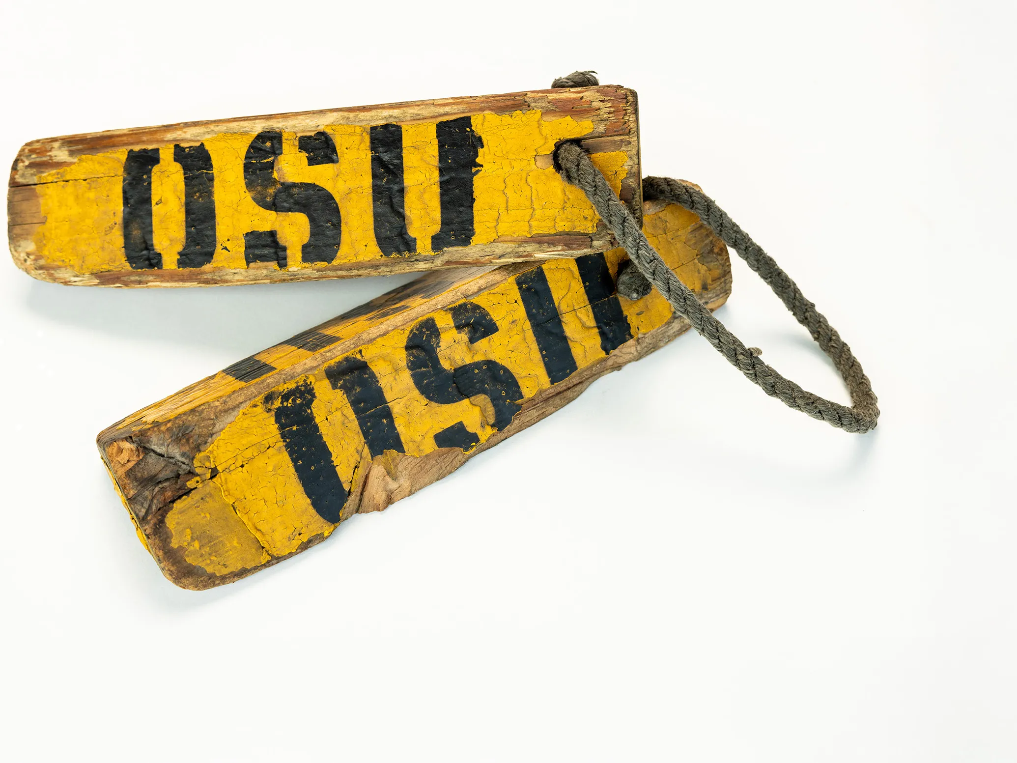 Two weathered, long triangles are tied together by a rope. They have the letters OSU stenciled on them and are shaped like this so that they can be placed on either side of an airplane wheel to stop it from rolling.