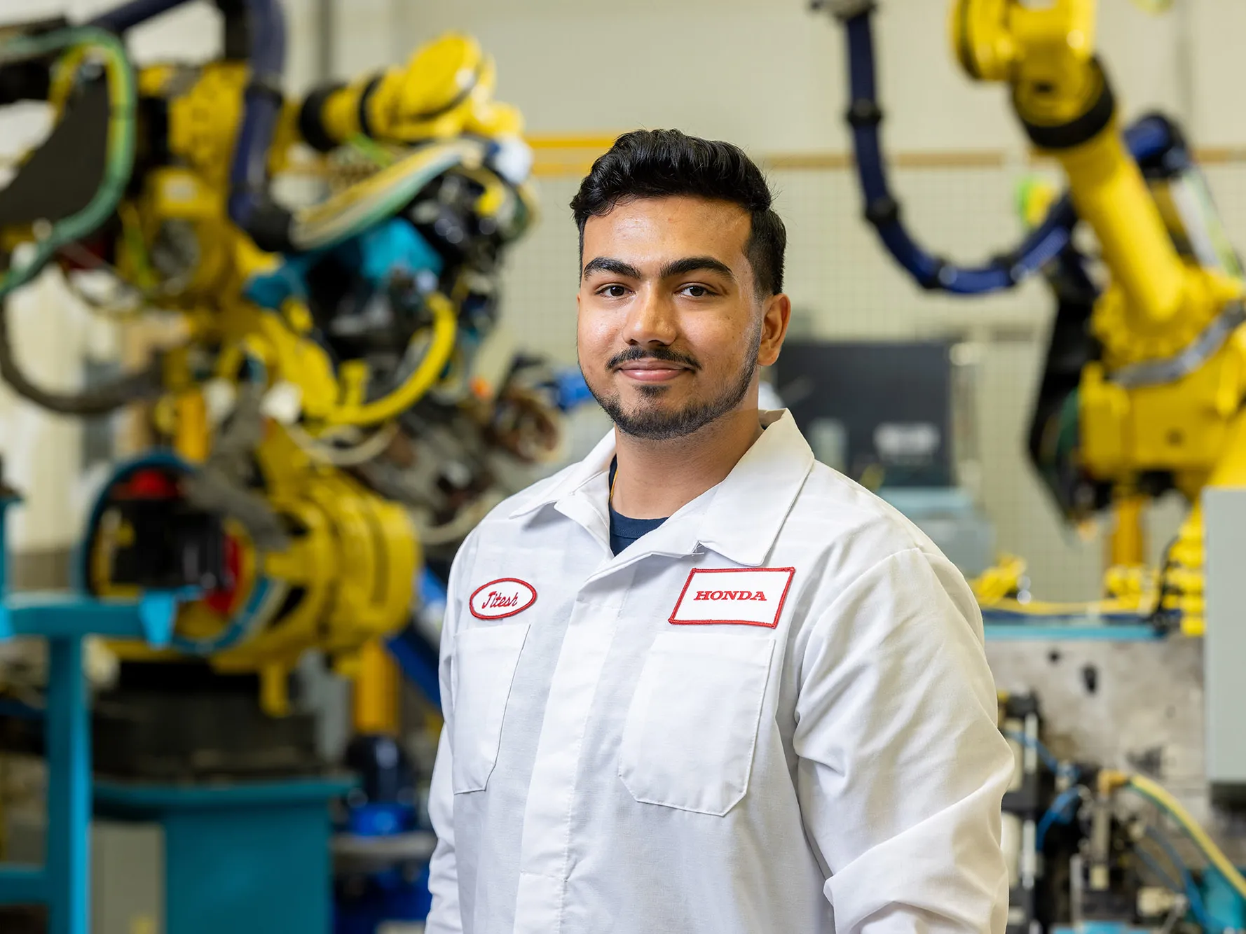 Jitesh Vidhani poses for a photo wearing his Honda uniform and in a hightech factory. Yellow, heavy duty robotic arms and other machinery is working in the background. Jitesh is slightly smiling, with his eyes and mouth.