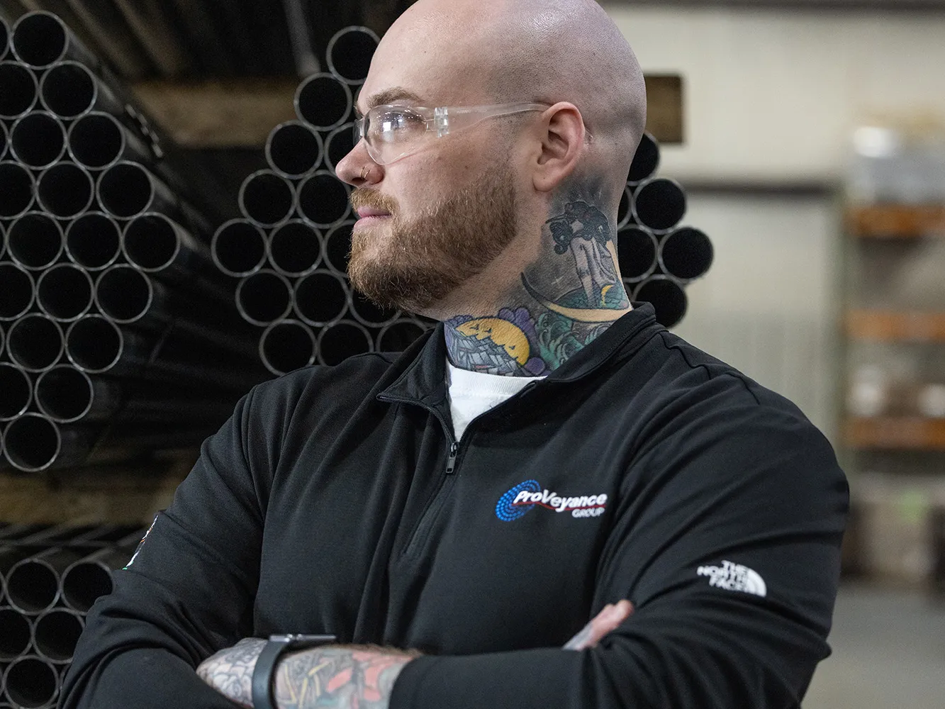 Zachary Ernest, a tall white man with shaved head, neat beard and colorful tattoos on his neck and forearms, is pleased by what he sees on the factory floor of his employer, Ashland Conveyor Products. He stands in front of bundles of about 50 metal pipes each, stacked on shelving. He also wears safety glasses.