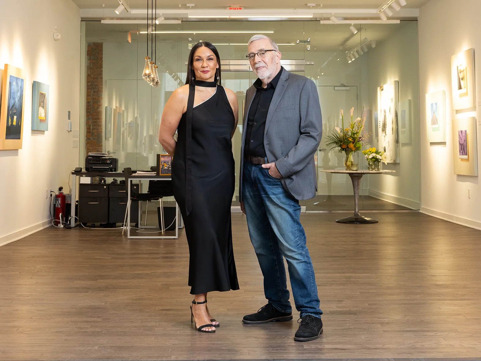 Michelle Brandt wears a black dress and Duff Lindsay wears jeans and a sports coat in a new art gallery. They seem elegant and thoughtful as they pose standing next to each other and slightly smiling.