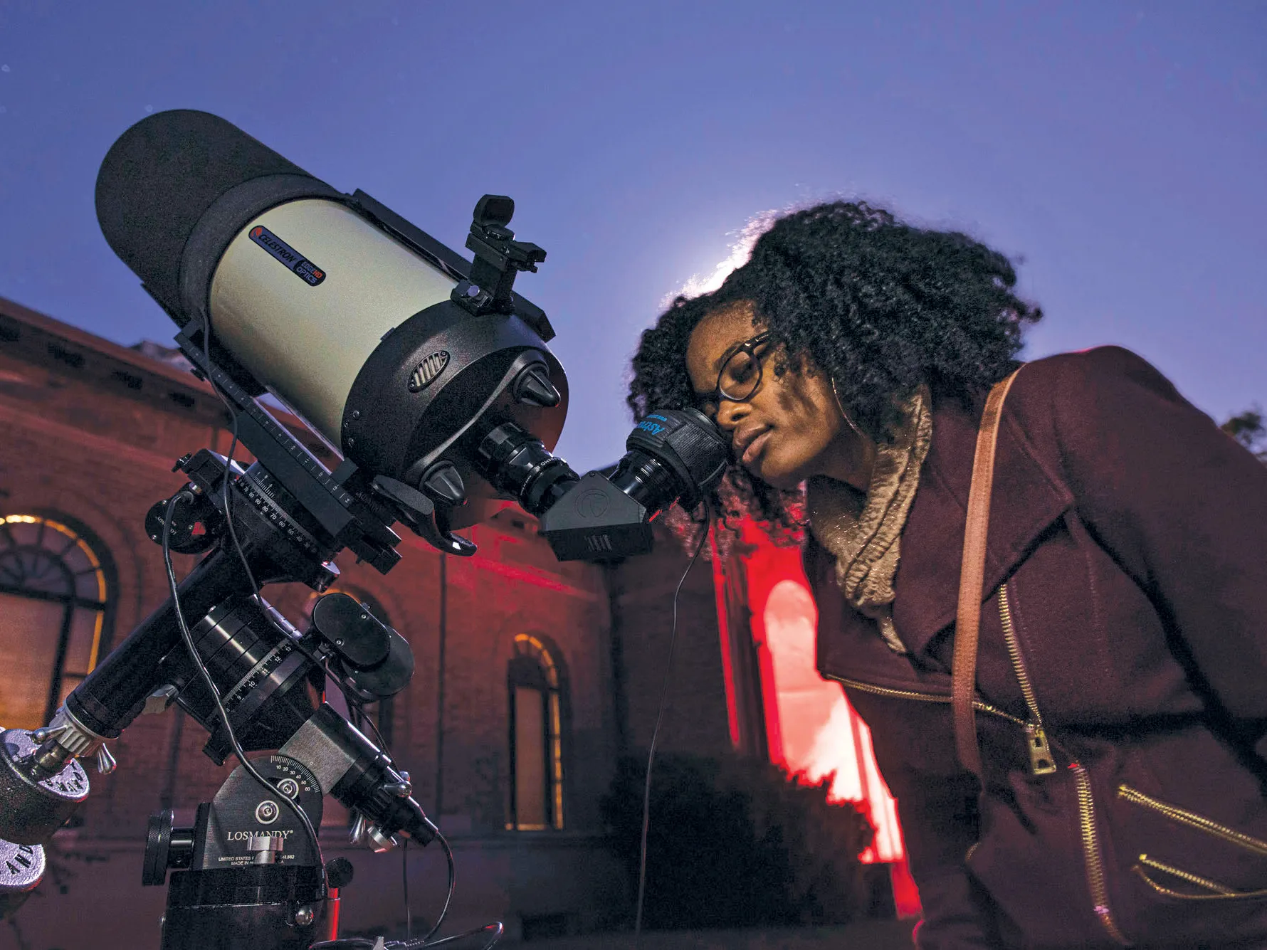 Caprice Phillips, a young black woman wearing her hair curly, a fall coat and glasses, peers into the scope of a telescope. Behind her the sky appears purple
