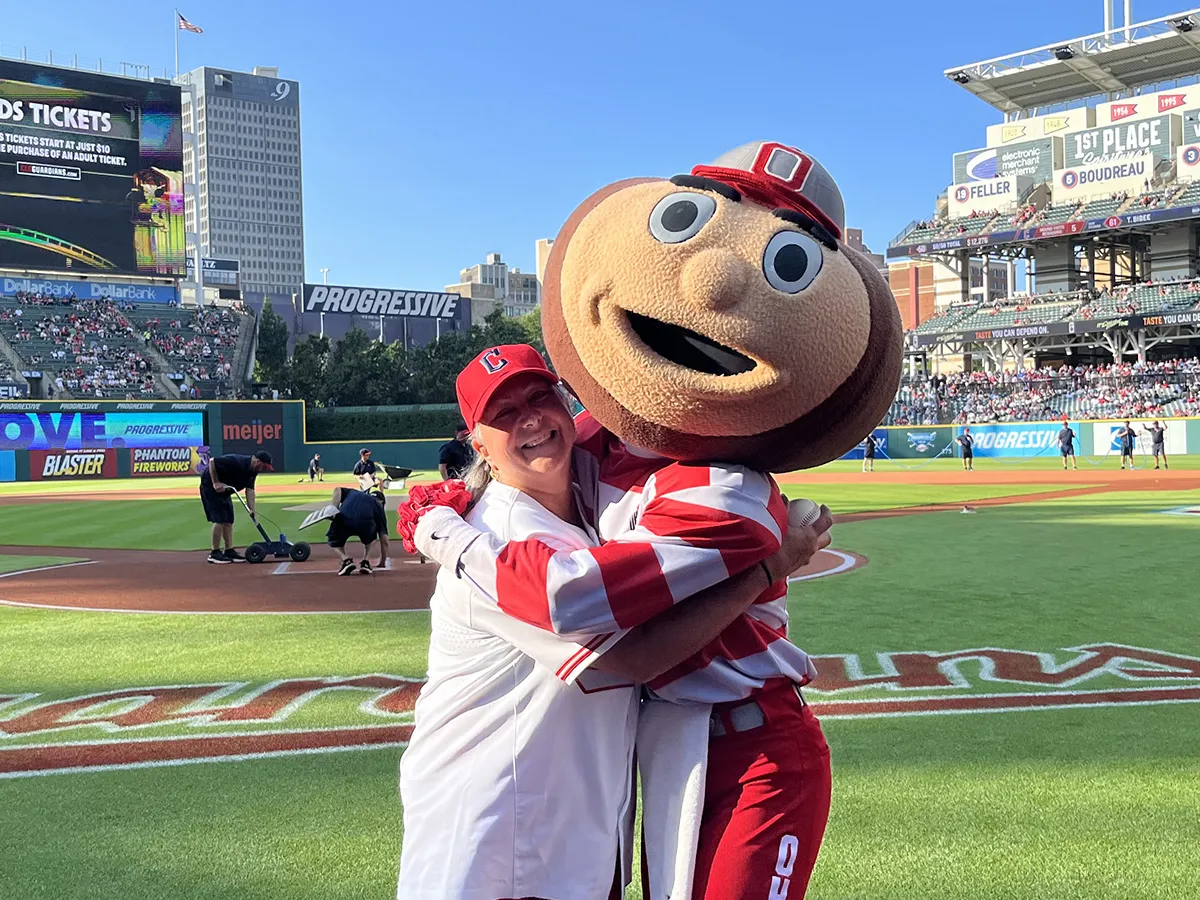 Molly Ranz Calhoun, wearing a baseball cap, T-shirt and shorts, laughs as she gives Brutus Buckeye, Ohio State&#039;s mascot with a buckeye-shaped giant head, a hug at the Cleveland Guardians&#039; baseball stadium. Molly is a middle-age white woman with shoulder-length hair and a friendly smile.