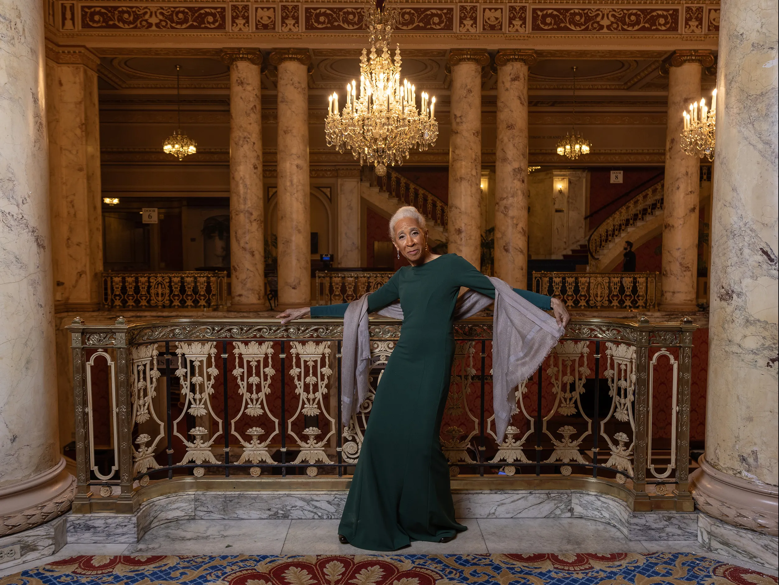 Dianne McIntyre, a slender and stylish black woman, poses along an ornate railing in a historical theatre with big marble columns and intricate detailing. Dianne is an attractive older woman. She has white hair pulled back in a bun, long legs and her body is curved as if in motion, with her arms outstretched. She wears a sleek column dress and one hand holds the end of a silk wrap draped around her back.