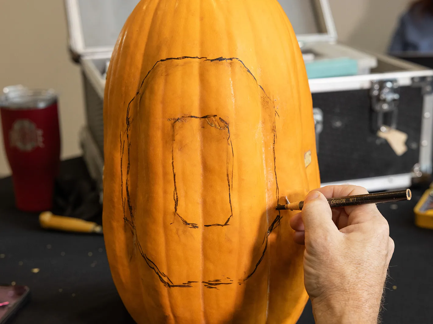Gu uses a crayon that looks more like a colored pencil to draw a Block O on the face of his tall pumpkin. Behind it on the table is his chest of tools and an insulated Ohio State cup.