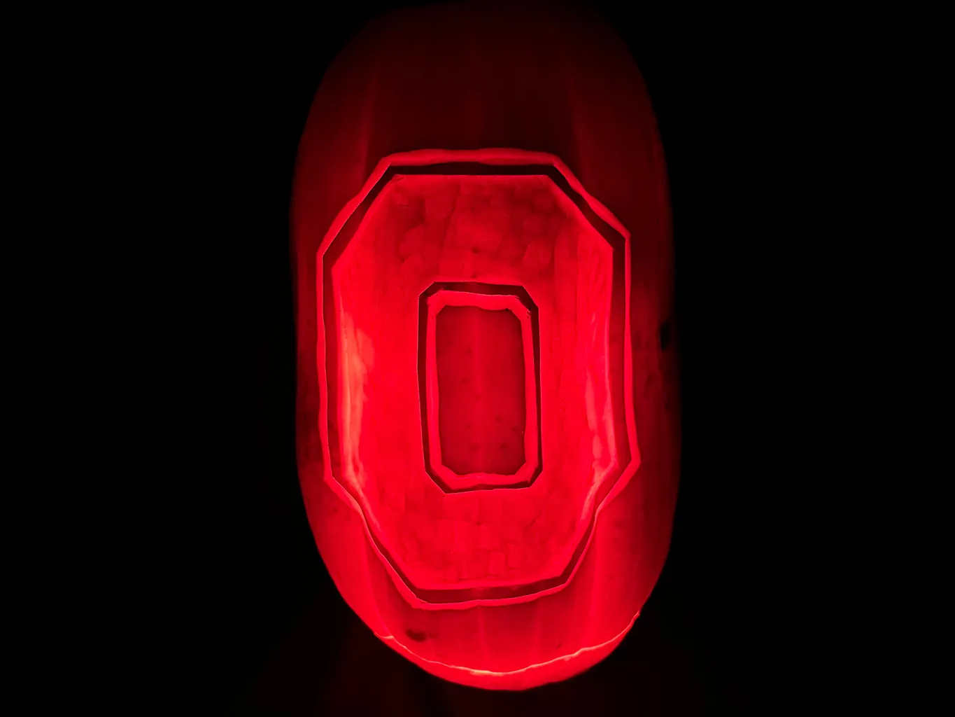 In a photo taken in the dark, a red-shining pumpkin emerges from the black. The Ohio State Block O appears super-clearly.
