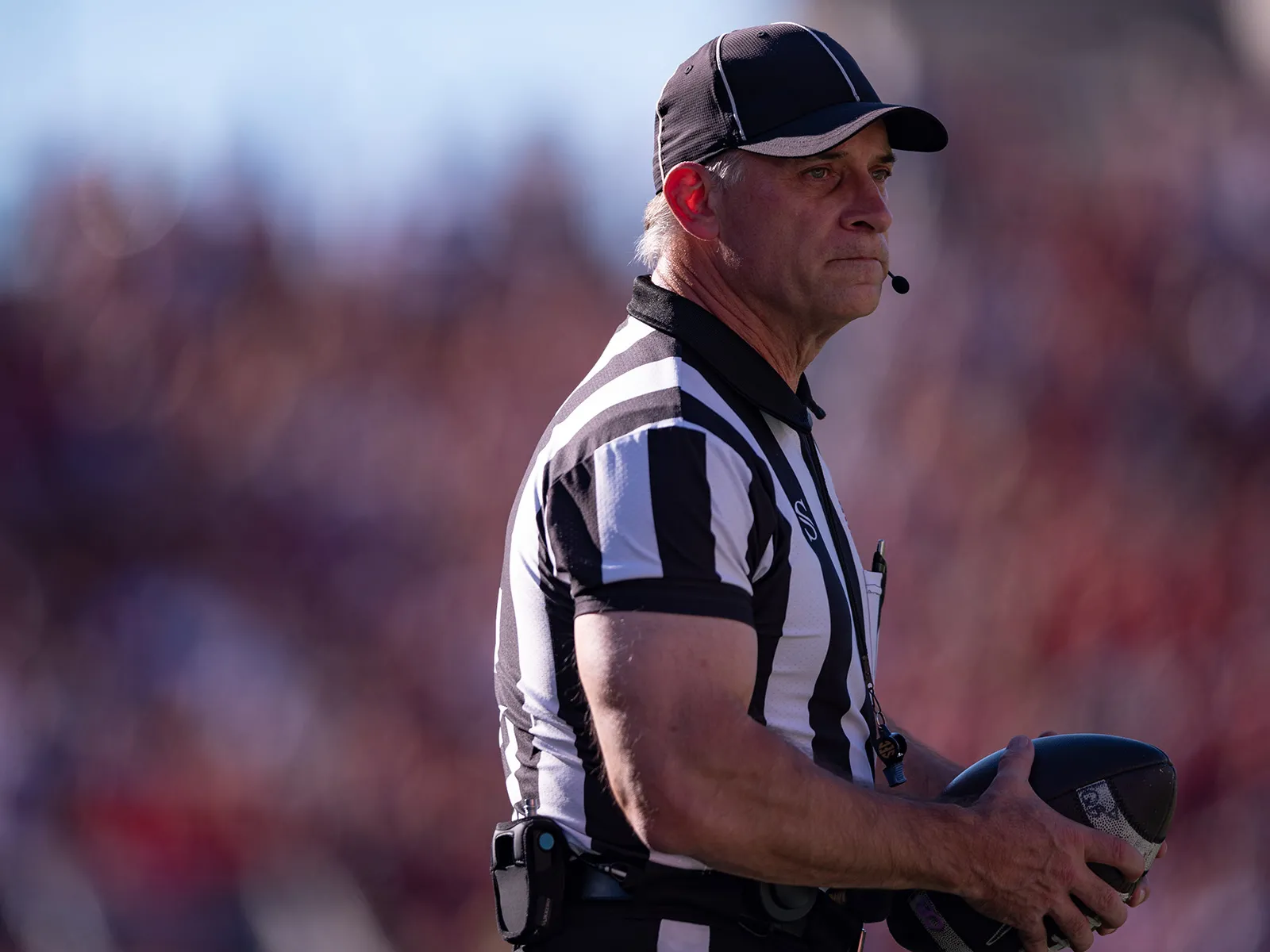 A football referee holds the ball and makes a thoughtful face as he watches something out of the photo frame. He wears a black and white striped referee’s shirt, a ballcap and a headset that leaves a small mic in front of his mouth to communicate calls. He&#039;s a white man with white hair and muscular arms. 