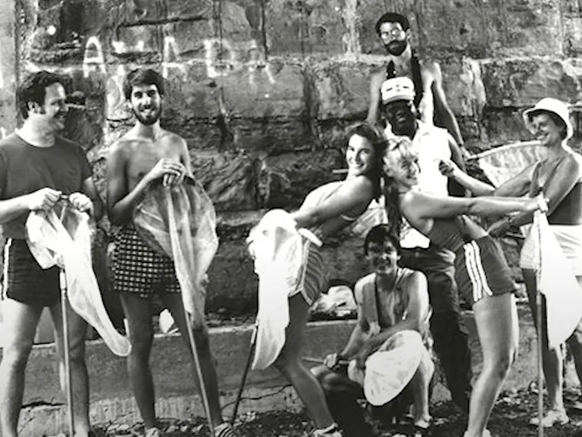 A group of students in swimwear clown around in front of a stone wall along the edge of Lake Erie