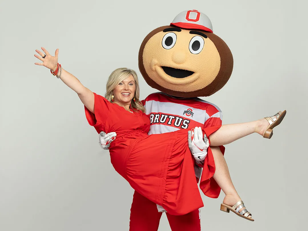 Tracy Stuck, a blond-haired white woman with a big smile and a scarlet dress, laughs as Brutus Buckeye hoists her in a cradle hold.