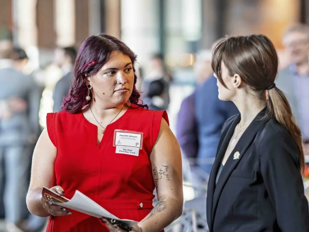 Danielle Johnson wears a scarlet dress and a focused expression as she talks to another student leader at an Ohio State hospitality event they coordinated. People dressed in suits can be seen in the background.