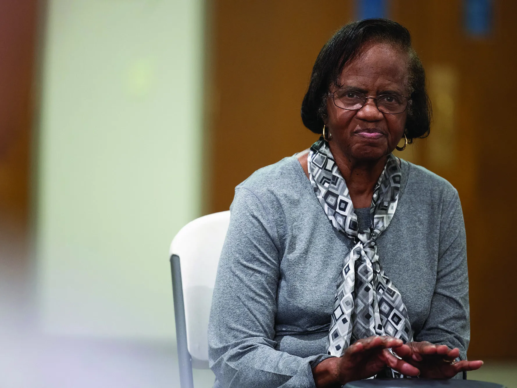 Ruthie Clemons, an older black woman with Parkinson&#039;s, stares into the camera as her portrait is taken. Her hands are poised over a drum. She&#039;s well dressed, wearing a pretty scarf, glasses and hoop earrings.