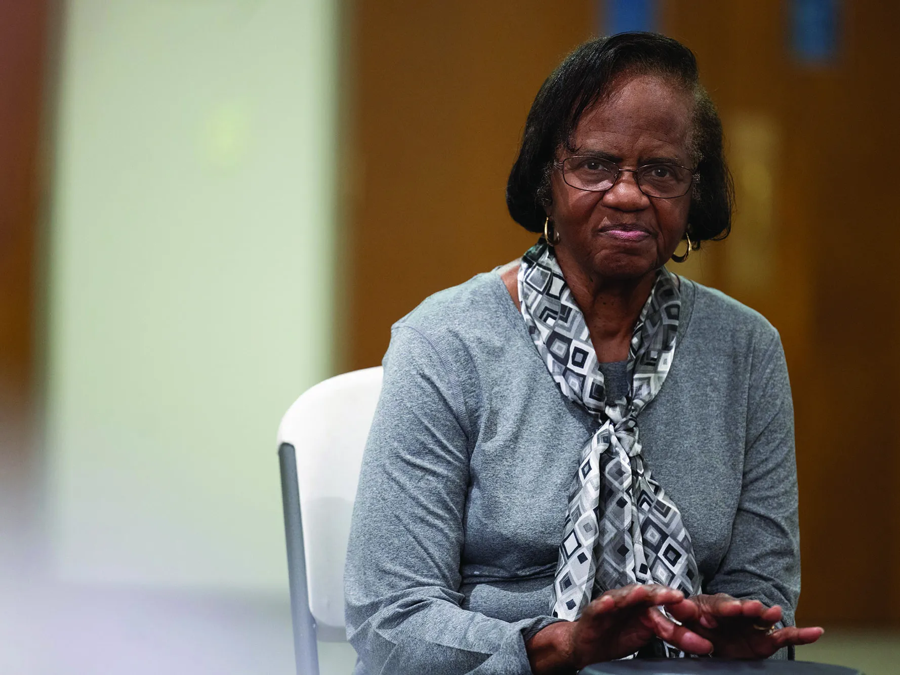 Ruthie Clemons, an older black woman with Parkinson's, stares into the camera as her portrait is taken. Her hands are poised over a drum. She's well dressed, wearing a pretty scarf, glasses and hoop earrings.