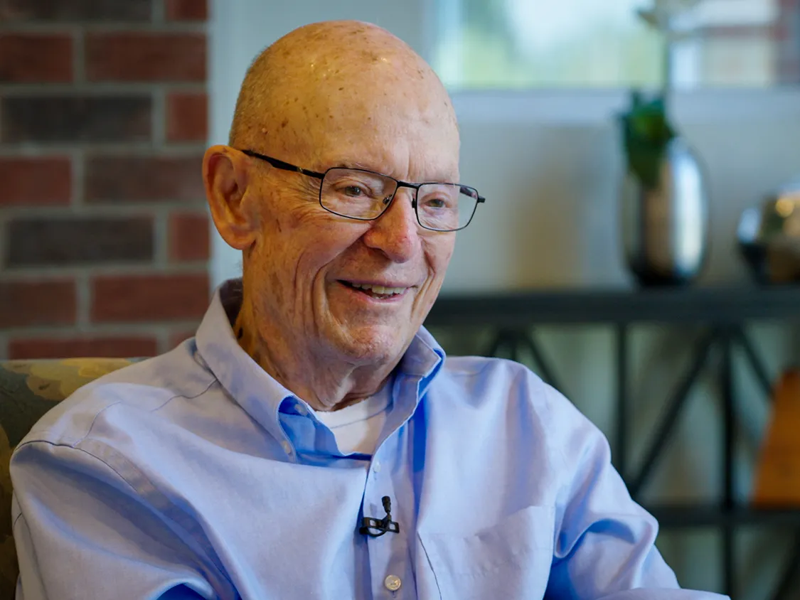 Dick Wood smiles as he tells a story. He&#039;s an older white man wearing glasses and a button-up shirt. Lines on his face shows he&#039;s smiled a lot through his years.