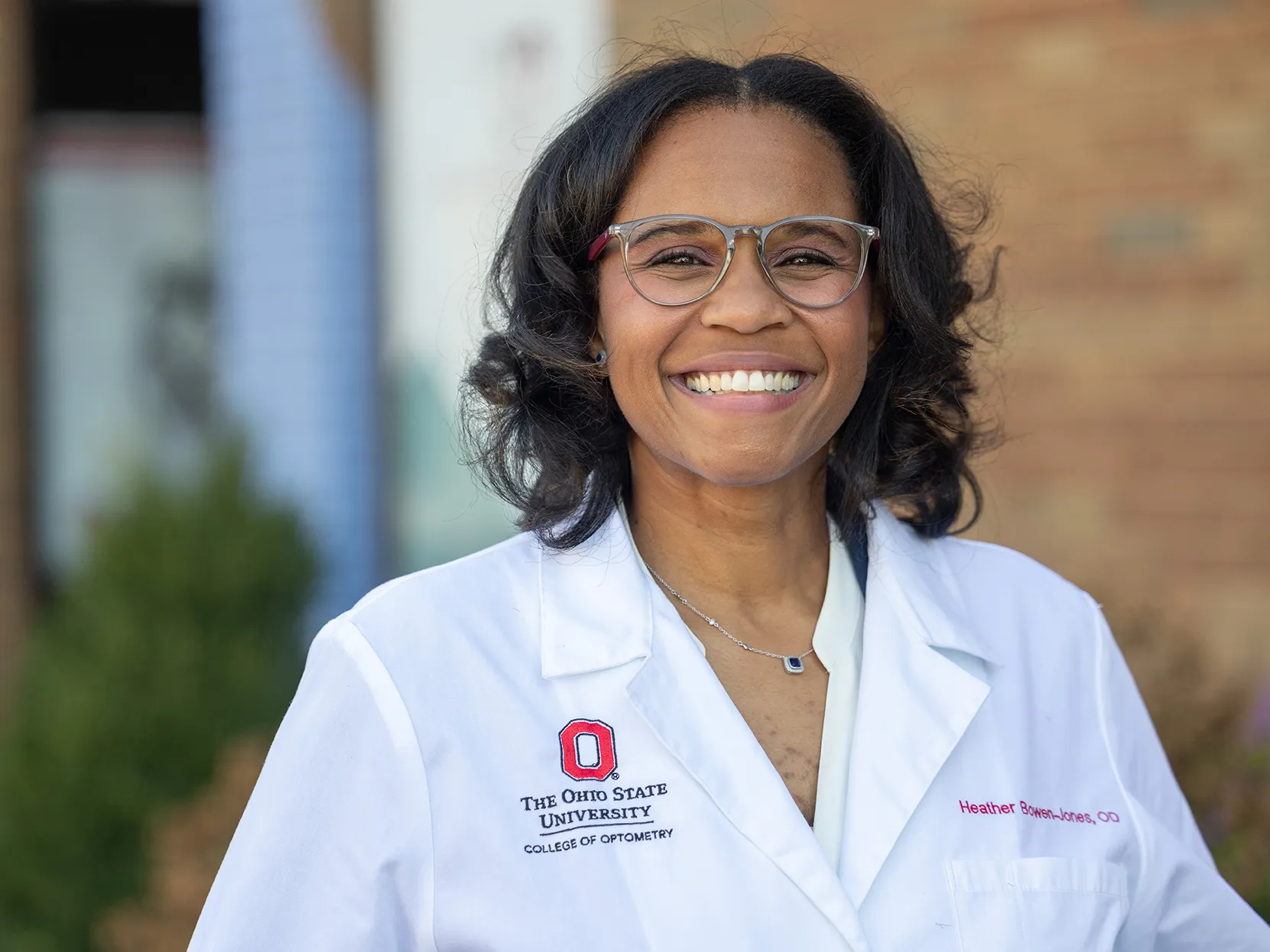 Heather Bowen-Jones ’11 OD grins for the photographer as she is photographed for a portrait. She stands in front of a medical building, wears glasses and a white doctor&#039;s coat, and she looks genuinely happy to share what she does.