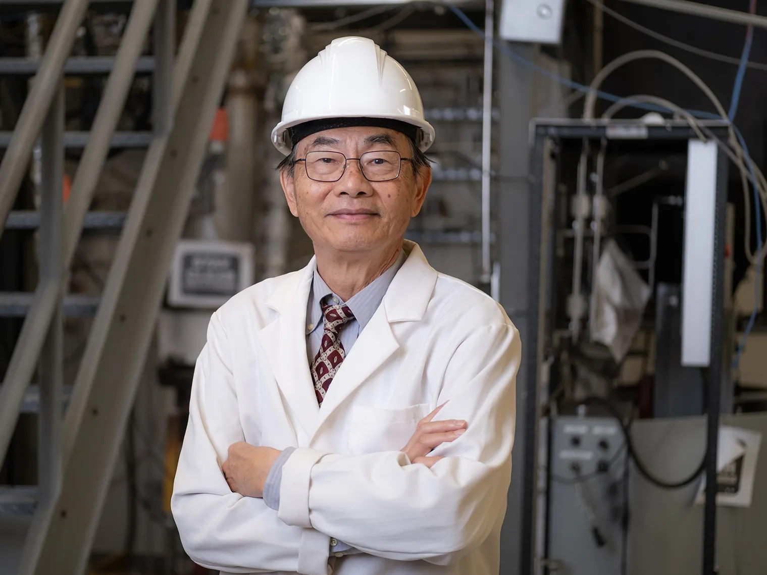 Liang-Shih Fan, an Asian man, seems quietly proud and confident, as he poses for this portrait. He wears round glasses and a white lab coat and hard hat over his suit. Behind him is the space where he works, filled with metal apparatus and tubing, as well as a staircase.