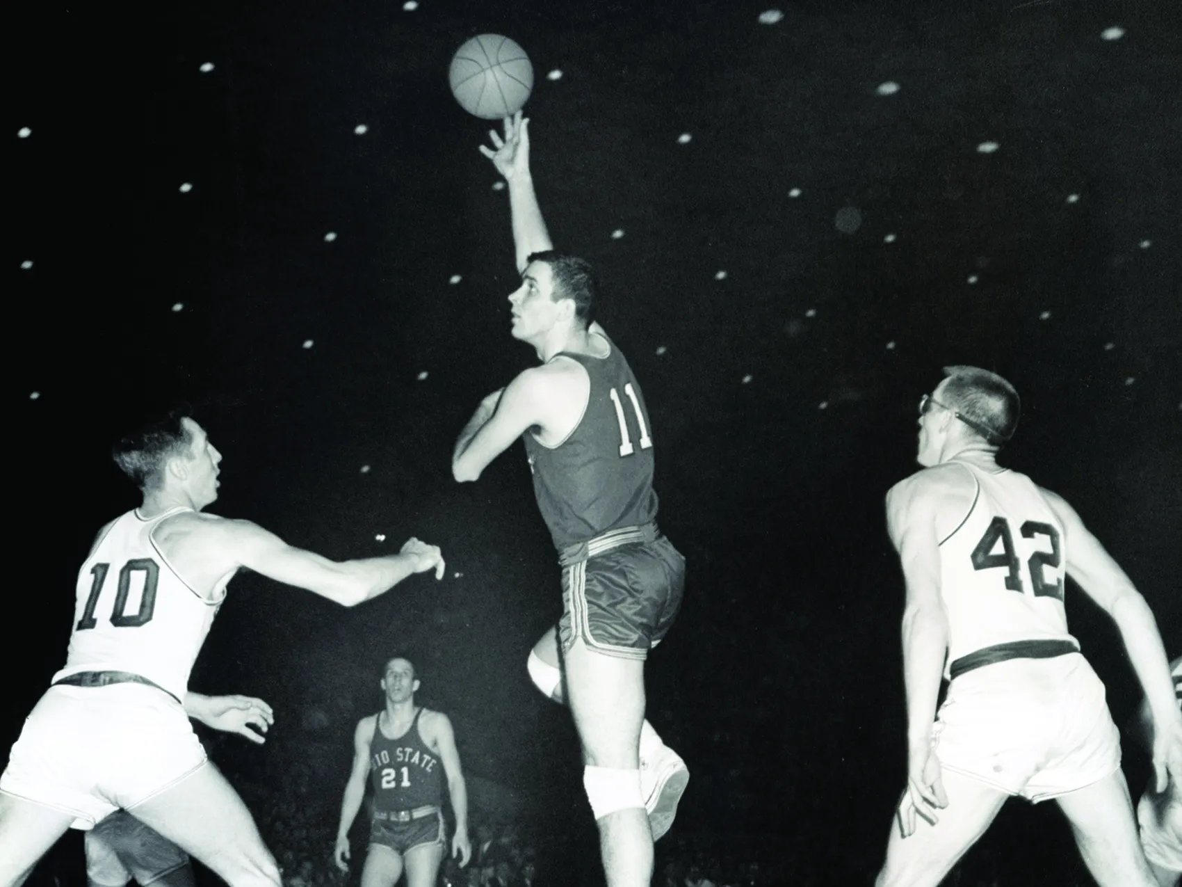 In an older black and white photo, an Ohio STate basketball olayer (Jerry Lucas) jumps high off the floor of the basketball court and hooks the ball over his head. Players from other teams look mini as he rises so high above them. His jersey is number 11 and he&#039;s. ayoung white man with neatly shorn hair and a strong build. He also wears knee pads.
