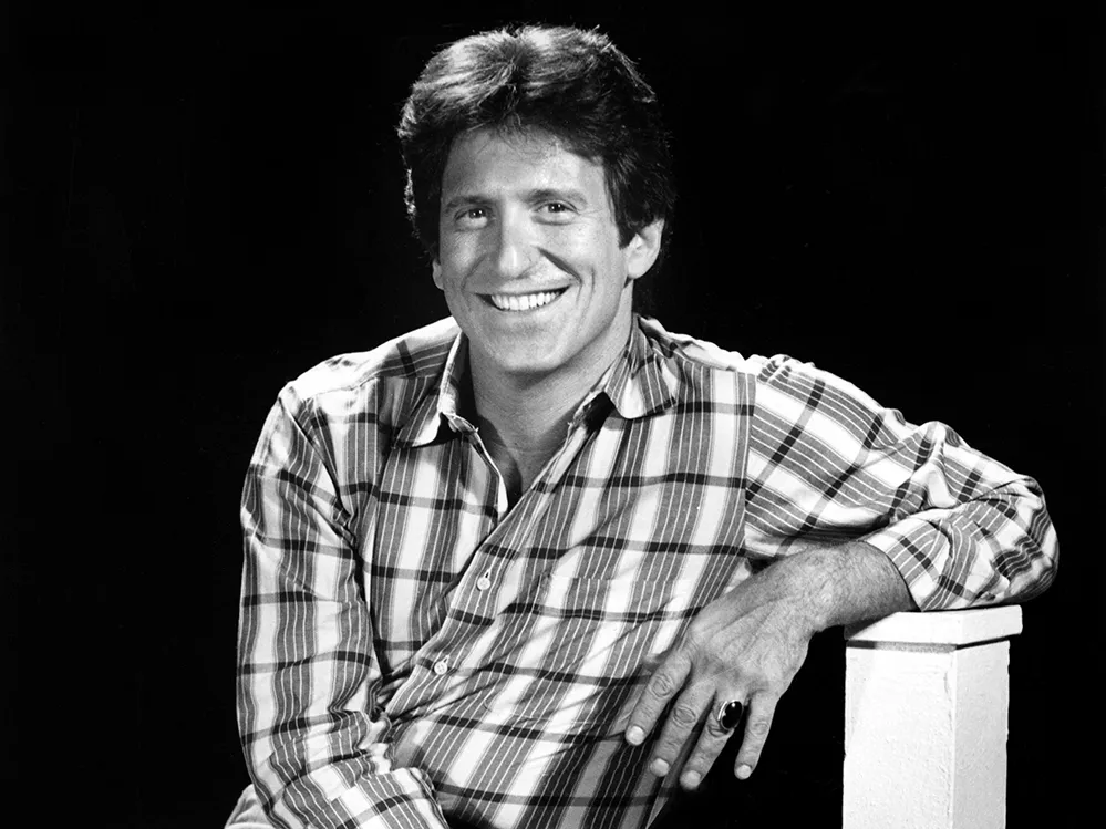 Michael Ochs smiles for a portrait as a younger man. A white man, he has thick hair, a big smile and wears a plaid shirt and jeans.