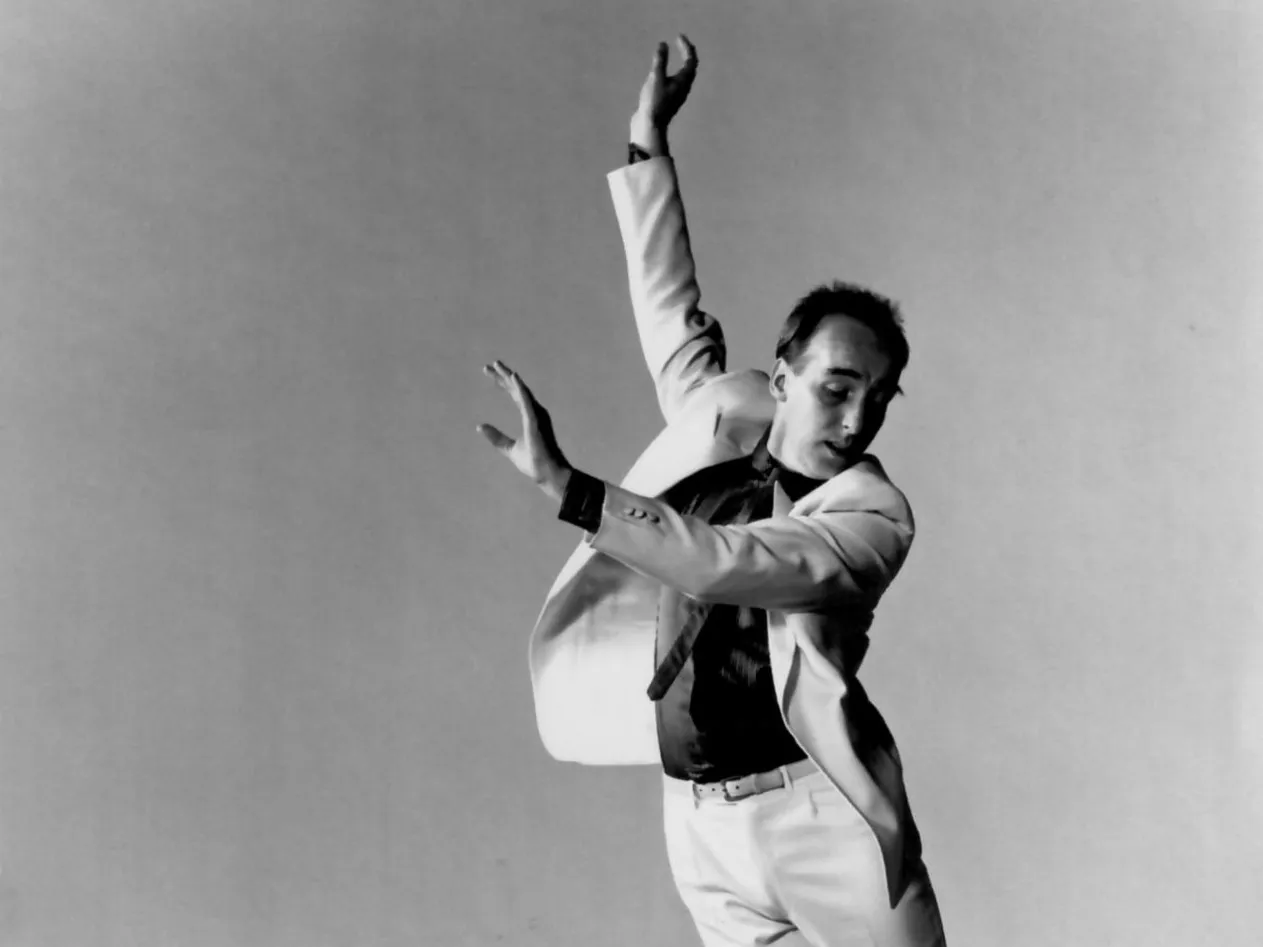 Fred Strickler tap dances with his full body slanted in a diagonal. He reaches up and out and looks down toward the stage.