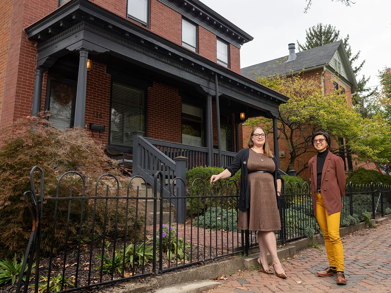 Two young people stand outside a well-kept brick duplex. They created the documentary on duplexes. One softly smiles and wears a dress, cardigan and black leather belt. The other smiles bigger and wears yellow pants and a brown suit jacket.