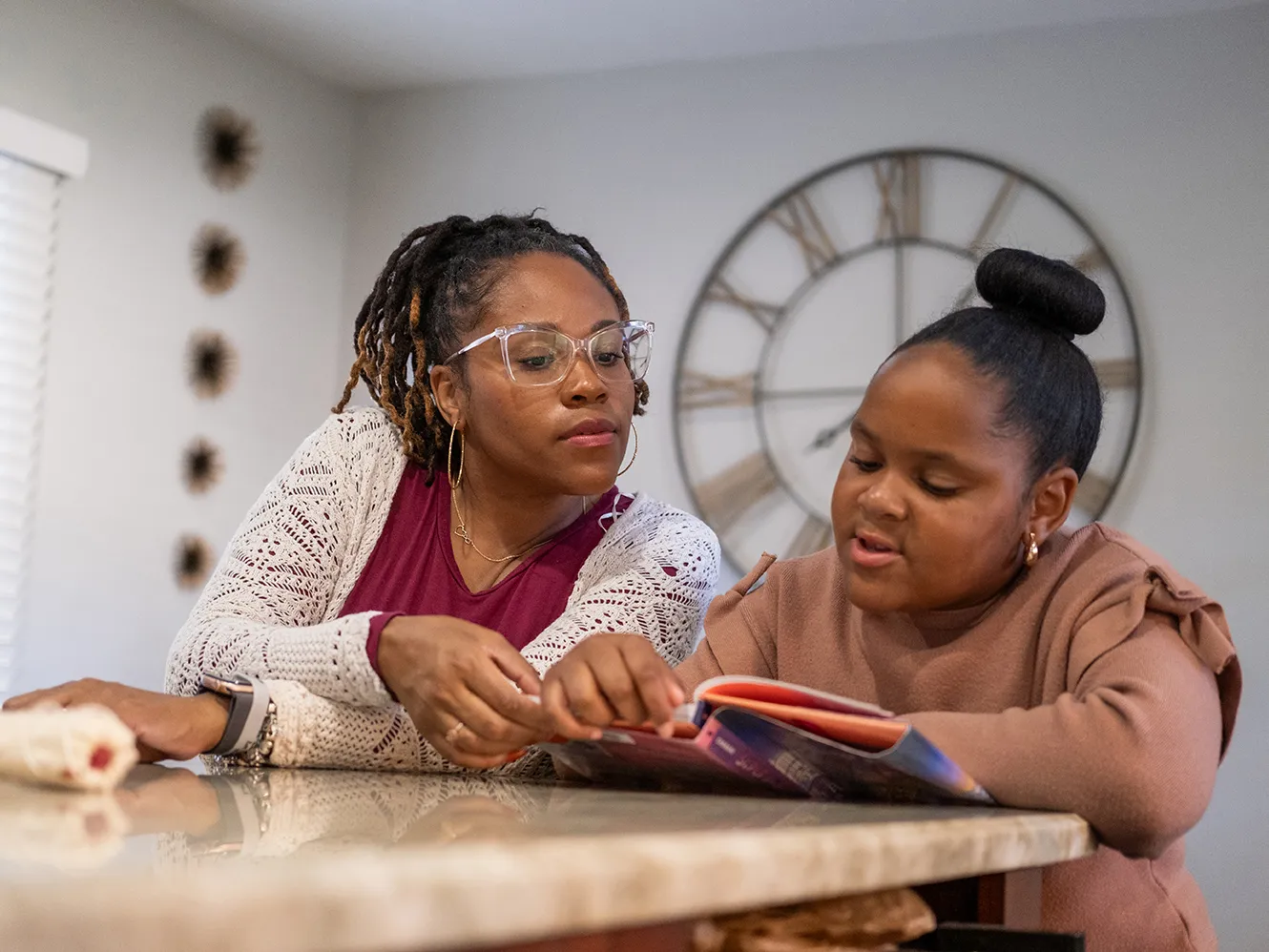 Nine-year-old Harper reads a thick children's book as her mom leans in to see the page. Harper is a cute Black girl with her hair pulled into a neat bun on top of her head, and her mom wears glasses and a pretty cardigan. They sit in their kitchen at home.