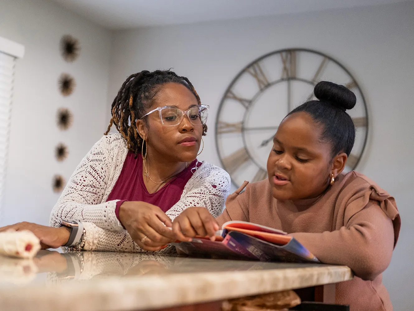 Nine-year-old Harper reads a thick children&#039;s book as her mom leans in to see the page. Harper is a cute Black girl with her hair pulled into a neat bun on top of her head, and her mom wears glasses and a pretty cardigan. They sit in their kitchen at home.