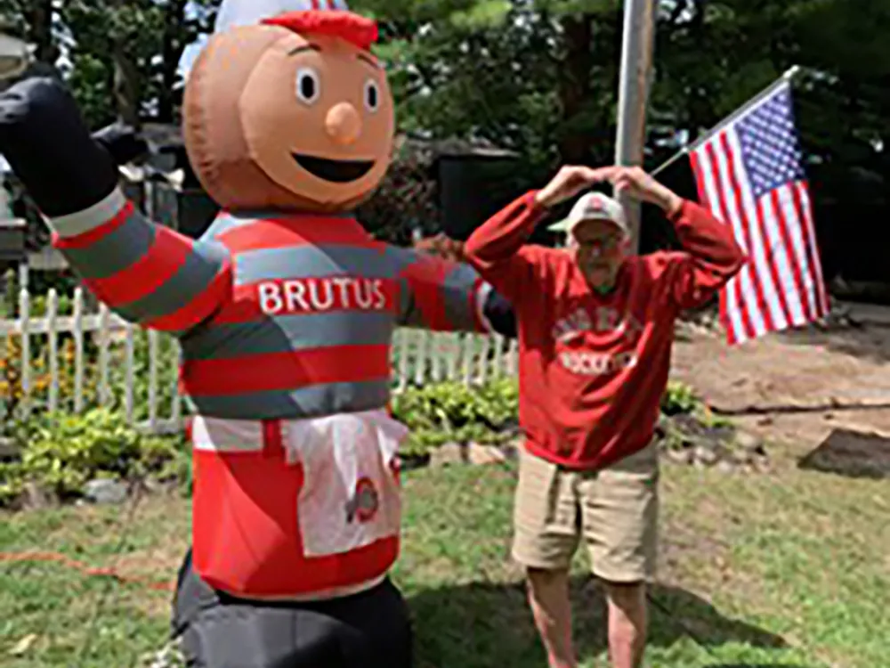 An older white man wearing an Ohio State sweatshirt makes a Block O standing next to a giant blow up Brutus Buckeye. It's maybe 150% as tall as he is. They're in a yard and an American flag flutters in the background.