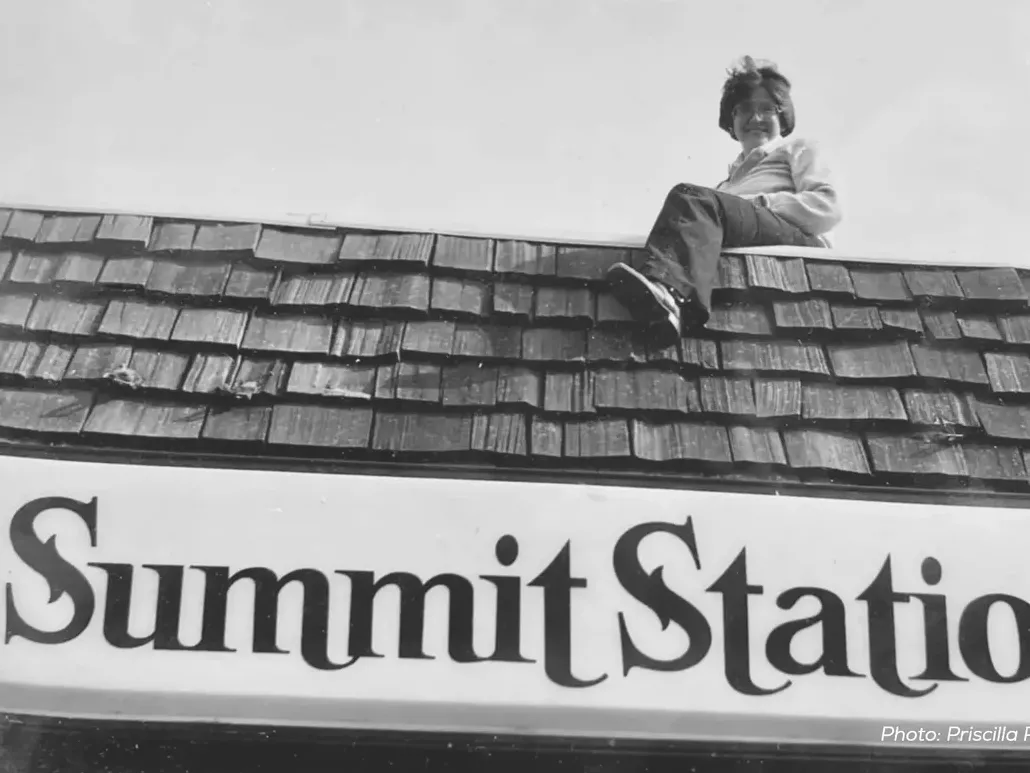 A woman grins as she sits on the roof of a bar. The sign beneath her says Summit Station.