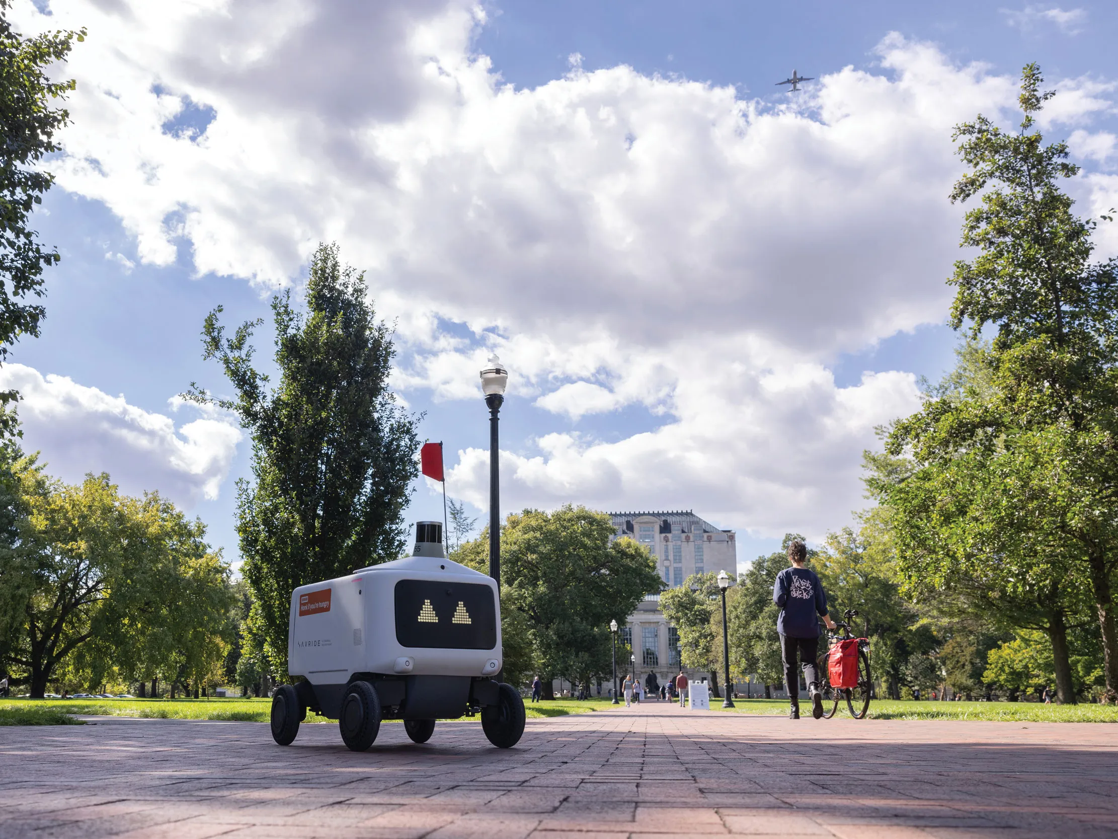 A lone food rover, which is a plastic-covered rectangle with a screen face showing trinagle-shape eyes, makes its way across the oval. Thompson Library and students can be seen in the distance.
